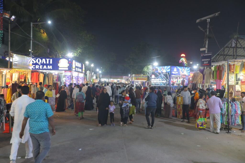 A crowded outdoor night market at Nampally Exhibition Grounds Hyderabad, with people walking and shopping at brightly lit stalls selling clothes and other items.